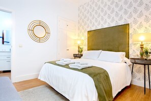 A neatly made bed with white linens and a green throw, two bedside lamps, and a wall with a leaf-patterned wallpaper.