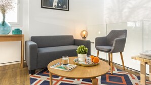 Un salon moderne avec un canapé gris, une table basse ronde en bois, un fauteuil gris, une petite plante en pot et un verre d’eau.