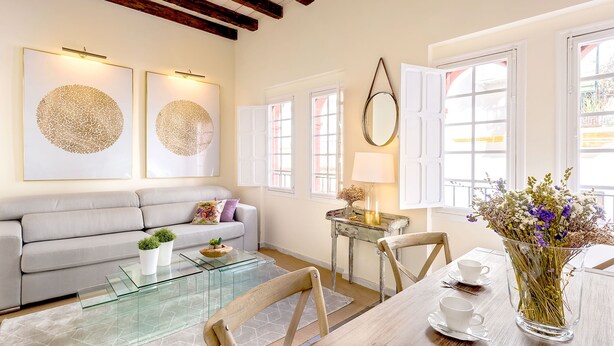 A modern living room with a sofa, glass coffee table, and a dining table with chairs. There are two windows with white shutters, a lamp on a side table, and artwork on the wall. - Genteel Home Galera Terrace (Seville)