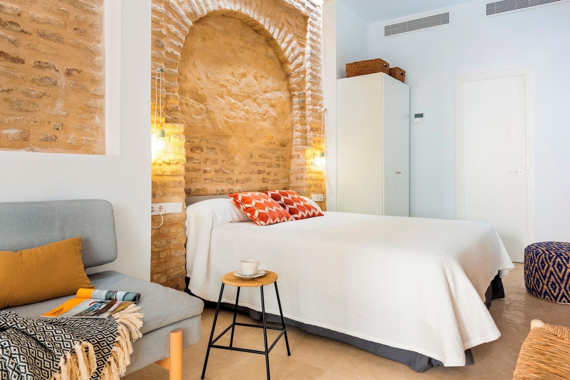 Un dormitorio acogedor con pared de ladrillo, cama con ropa de cama blanca, un sofá gris y un taburete pequeño.