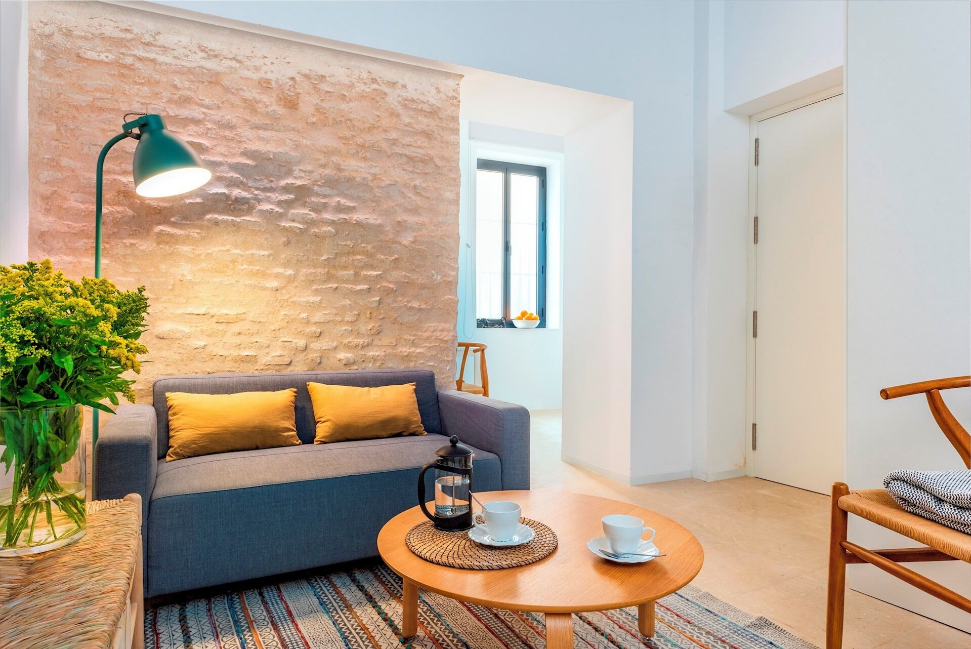 A cozy living room with a grey sofa, yellow cushions, a round wooden coffee table with cups, a green floor lamp, and a rustic brick wall.