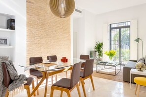 A modern living room with a glass dining table, comfortable seating, a brick accent wall, and a large window with a view. - Genteel Home Santa Teresa (Seville)