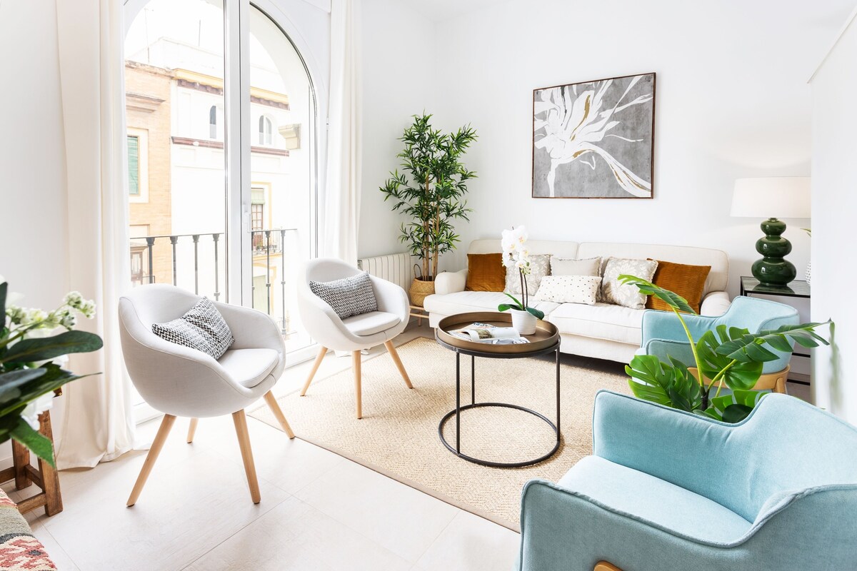 A modern living room with a white sofa, two armchairs, a coffee table, and a view of the city through large windows.