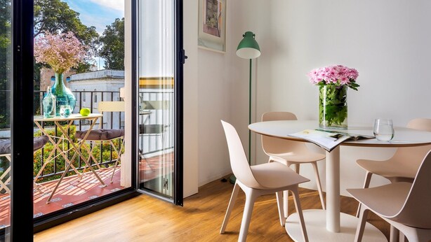 Une chambre moderne avec un balcon, une table à manger avec des chaises, un lampadaire vert et un vase rempli de fleurs roses.