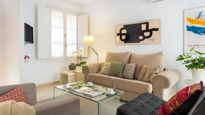 A modern living room with a beige sofa, a glass coffee table, and a black leather chair. There is a flat-screen TV mounted on the wall, a lamp, and a framed abstract painting. - Genteel Home Conde de Ibarra (Seville)