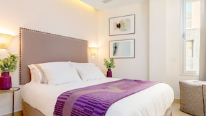 Ein ordentlich bezogenes Bett mit einer violetten Decke, einem Kopfteil, Nachttischlampen und zwei gerahmten Kunstwerken an der Wand.