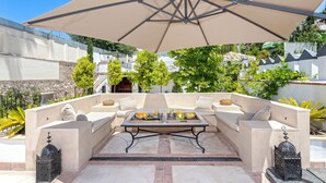 Un área de estar al aire libre con un sofá color beige, una mesa con frutas y bebidas, y una sombrilla grande que proporciona sombra.