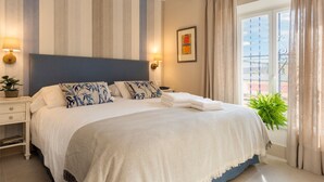 A neatly made bed with a white and blue patterned pillow set, a grey headboard, and a beige throw blanket. There is a small bedside table with a lamp and a plant. A framed picture hangs on the wall, and a window with curtains is visible. - Genteel Home Carmen San Ignacio I (Granada)