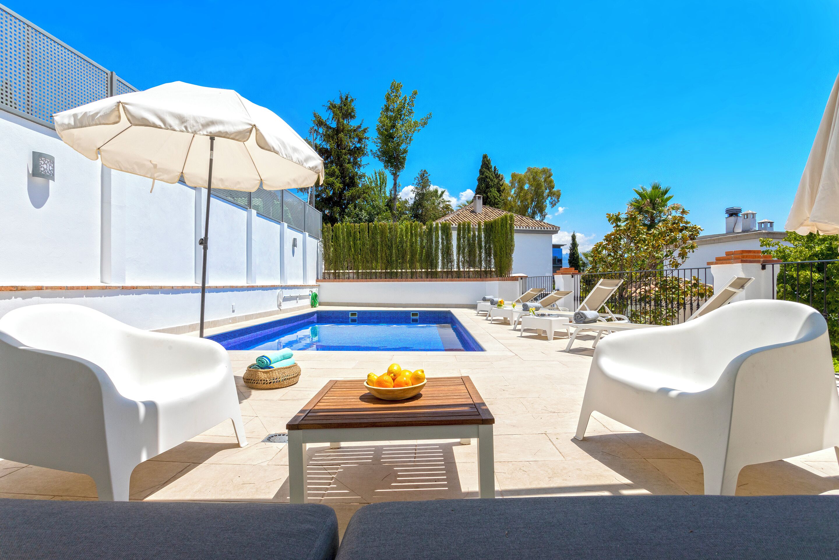 A poolside area with a wooden table, white chairs, and a white umbrella.