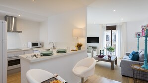 A modern kitchen with white cabinets, a microwave, and a stove. There is a dining area with a white chair and a table, and a living room with a sofa and a TV. - Genteel Home Cabildo III (Seville)