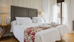 A double bed with a grey upholstered headboard, white bedding, and a floral-patterned throw. There are two bedside tables with lamps, a window with sheer curtains, and a view of the outdoors. - Genteel Home Cabildo III (Seville)