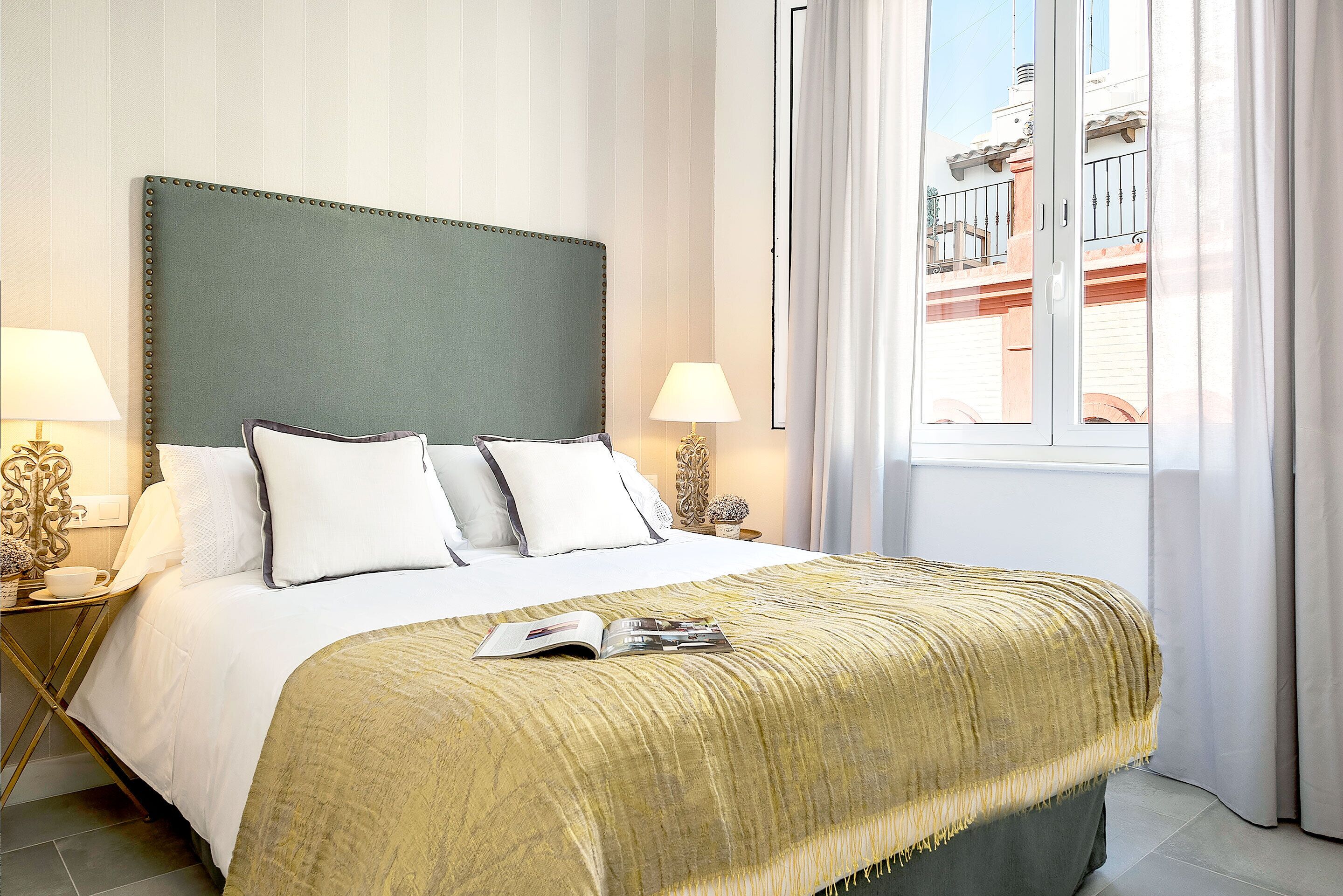 Una cama bien hecha con cabecera verde, ropa de cama blanca y una manta amarilla. Hay dos lámparas de noche y una ventana con cortinas.