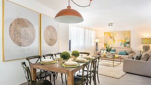 A dining area with a wooden table, chairs, and a pendant light. There is a living area with a sofa and a painting on the wall. - Genteel Home Plaza de las Flores (Málaga)