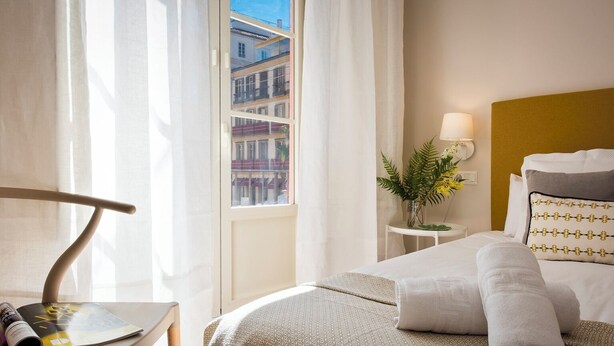 Habitació d'hotel amb un llit, una taula de nit amb una làmpada, una finestra amb cortines i vista a edificis.