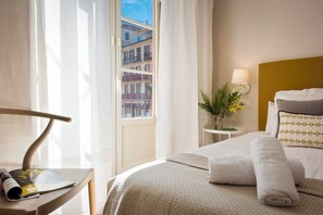 A hotel room with a bed, a bedside table with a lamp, a window with curtains, and a view of buildings outside. - Genteel Home Picasso I (Málaga)