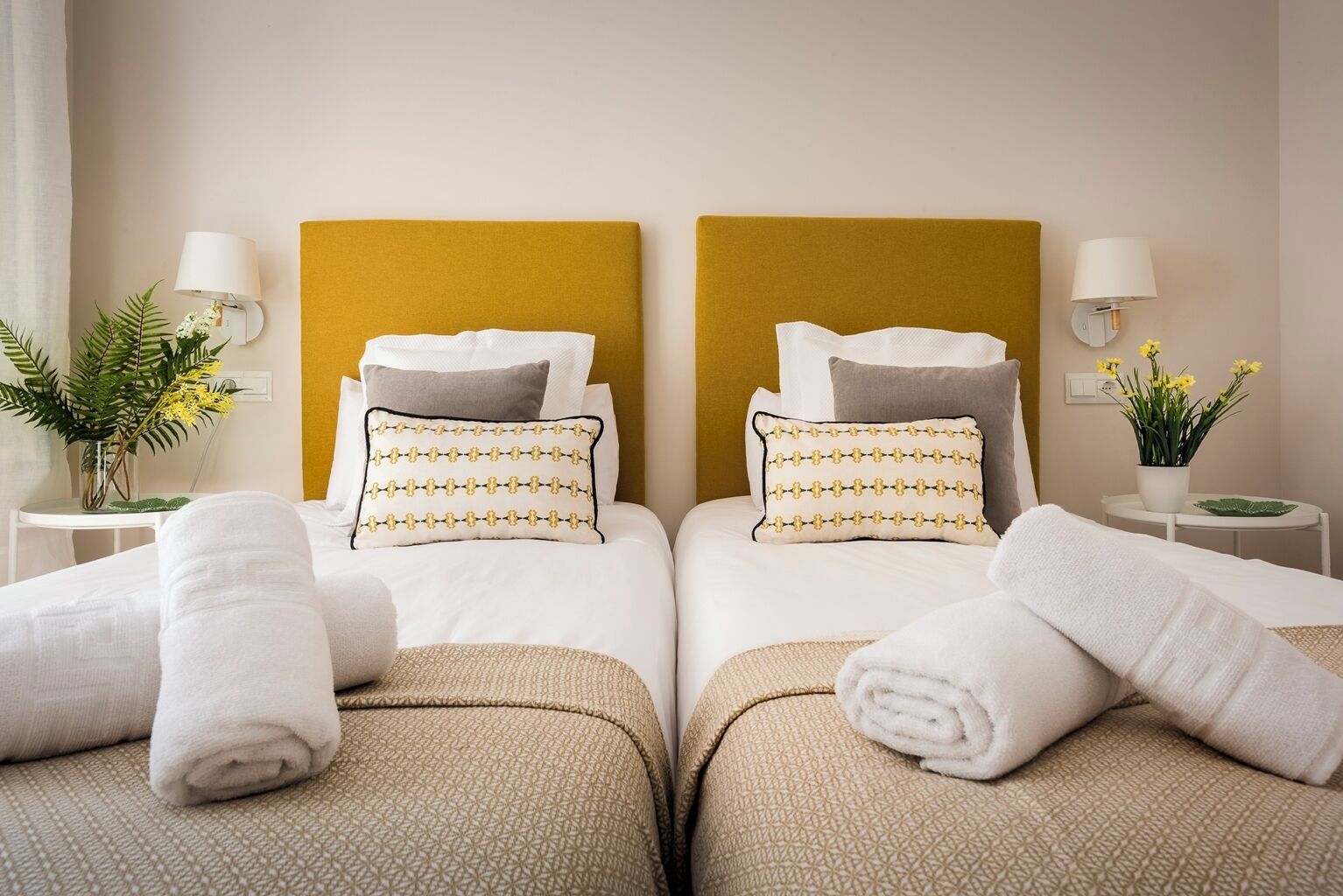 Hotellihuoneessa on kaksi sänkyä, joissa on molemmissa pääty sekä valkoiset vuodevaatteet ja kuviolliset tyynyt. Huoneessa on valkoiset yöpöydät, joilla on lamput ja pieniä ruukkukasveja.