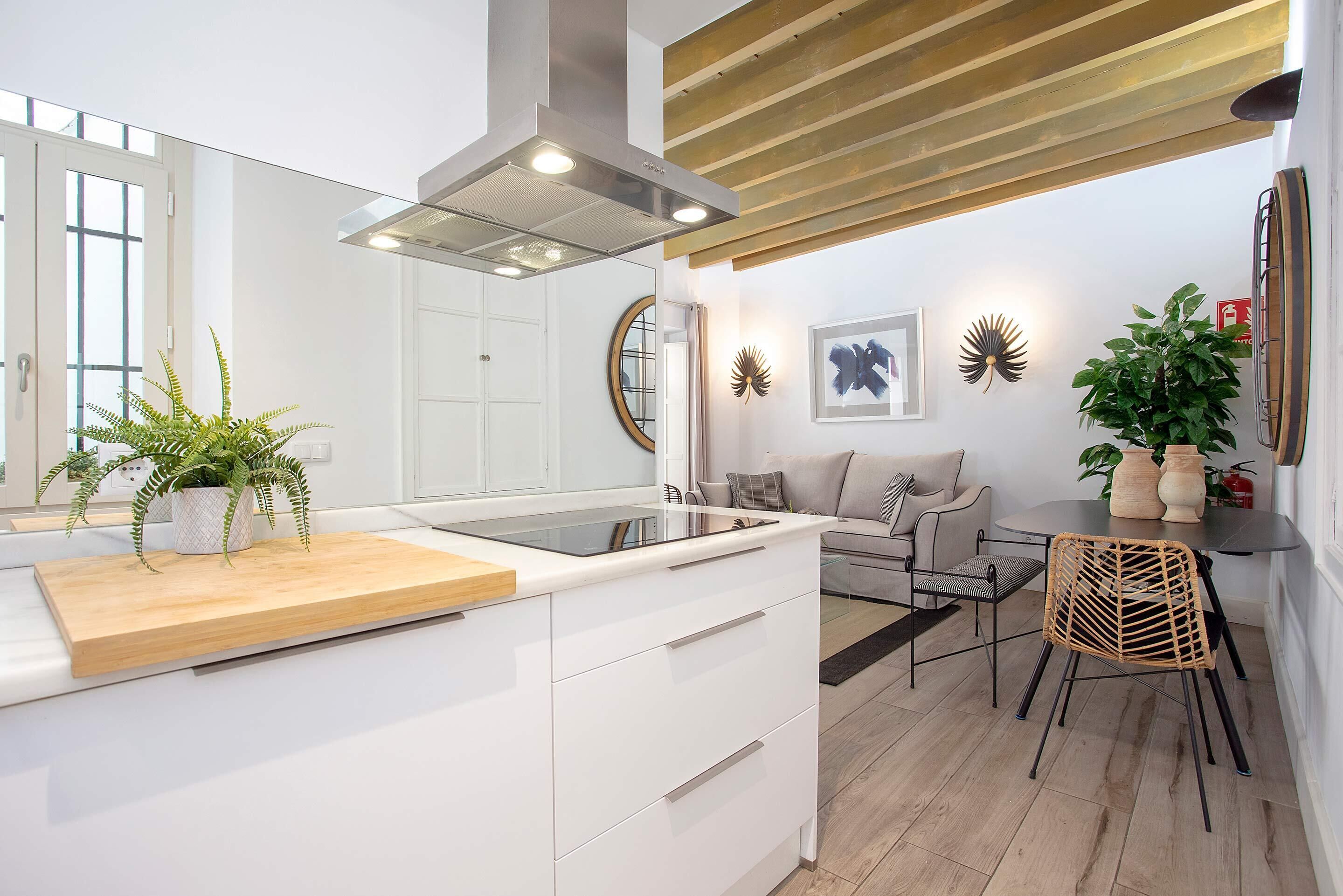 A modern kitchen with a wooden countertop, white cabinetry, and a central island. There is a dining area with a round table and chairs, and a sofa in the background.