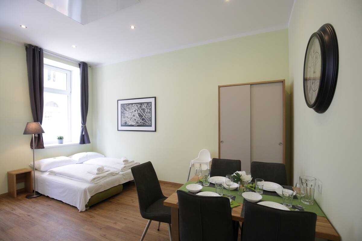 A hotel room with a bed, a dining table set for two, a chair, a bedside table with a lamp, a framed picture on the wall, and a clock.