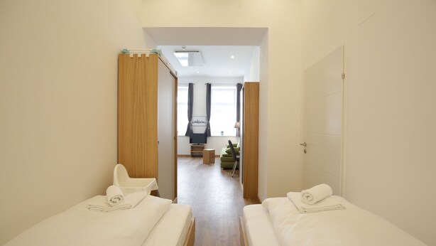 Quarto de hotel com duas camas de solteiro, um guarda-roupa de madeira, um pequeno espaço de estar e vista para o corredor.