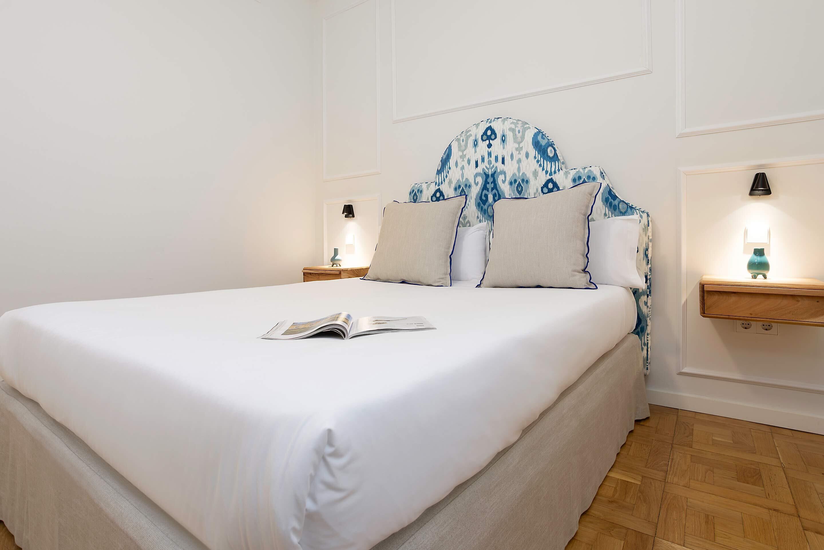 A neatly made bed with a white sheet, a pillow with a blue pattern, and a book on the bed. There are two bedside tables with lamps.