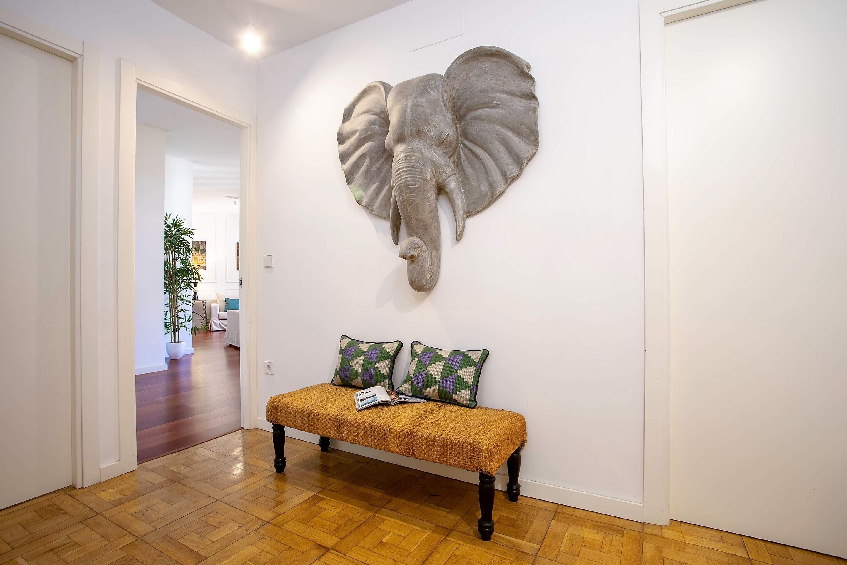 A hallway with a wooden floor, a bench with patterned cushions, and a large elephant head sculpture on the wall.