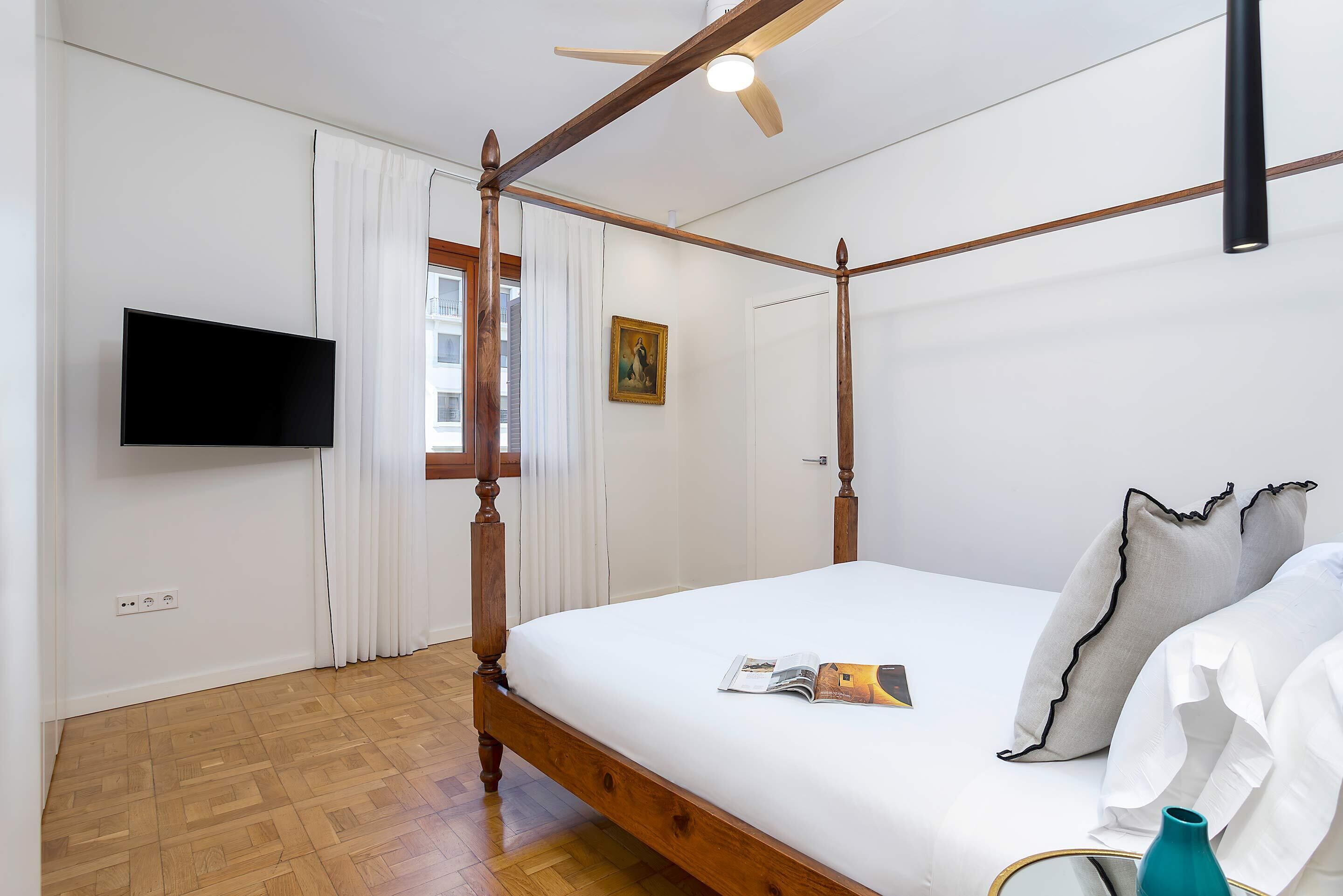 Un lit à baldaquin avec un téléviseur à écran plat fixé au mur, un ventilateur de plafond et un plancher en bois.