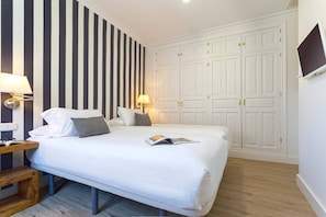 Une chambre d’hôtel avec un lit, des lampes de chevet, une table de nuit en bois, un magazine sur le lit, un téléviseur fixé au mur et une armoire à deux portes.