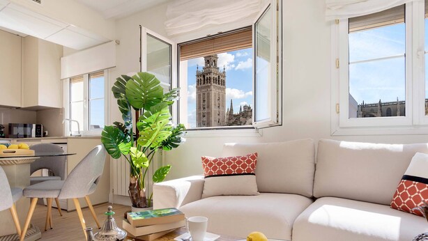 Una sala moderna con un sofá, una mesa de comedor con sillas, una mesa baja con una tetera y limones, y un ventanal con vistas a una torre histórica.