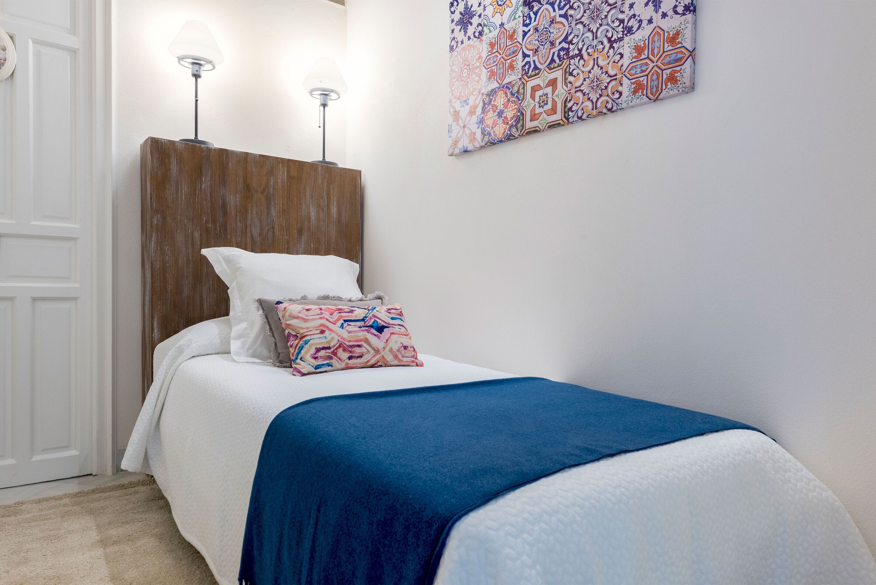 Un dormitorio con cabecera de madera, colcha blanca, manta azul y almohada colorida. Incluye una lámpara de pared y un elemento decorativo en la pared hecho de azulejos.