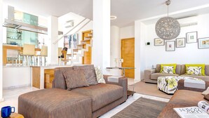A modern living room with a sofa, ottoman, and a coffee table. There is a staircase leading to an upper level, and a kitchen area is visible in the background. - Genteel Home Real de la Carreteria (Seville)