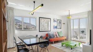A modern living room with a glass dining table, black chairs, a green bench, and a sofa with colorful cushions. There are framed artworks on the walls and a view of a cityscape through large windows. - Rooftop Funchal - By Wehost (Funchal)