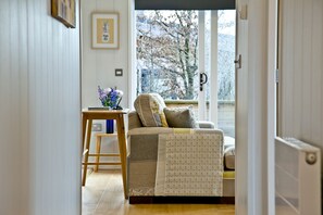 A cozy living room with a sofa, a small wooden table with a vase of flowers, and a view of a snowy landscape through a large window. - Nyland Lodge, Hot Tub - Fireplace- Free WiFi & Parking (Cheddar)