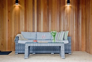 A wicker sofa with a grey cushion and striped pillows, a glass coffee table with a vase of flowers and two glasses of wine, and a black doormat on a wooden floor. - Nyland Lodge, Hot Tub - Fireplace- Free WiFi & Parking (Cheddar)