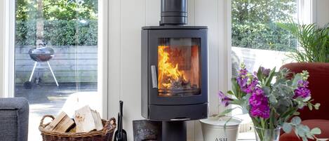 A modern living room with a wood-burning stove, a wicker basket with firewood, a vase with flowers, and a view of the outdoors.