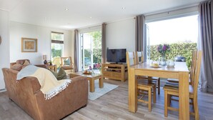 Een ruime woonkamer met een bruine bank, een houten eettafel met stoelen, een televisie en een groot raam met gordijnen.