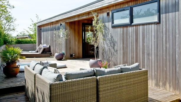 A modern building with a wooden exterior, large windows, and a patio area with wicker furniture and potted plants. - Mulberry Lodge, Strawberryfield Park (Cheddar)