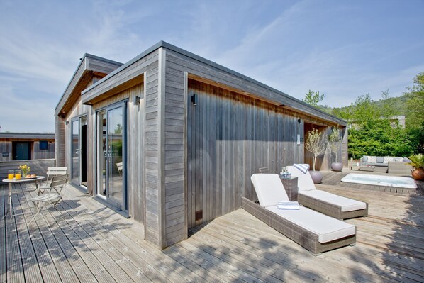 A modern outdoor deck with a wooden deck, a glass-enclosed patio area, and a lounge area with white cushioned loungers.