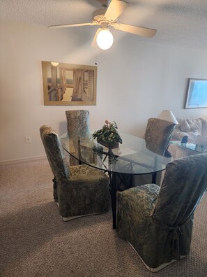Un coin repas avec une table en verre entourée de chaises à motifs, un ventilateur de plafond et un tableau encadré au mur.