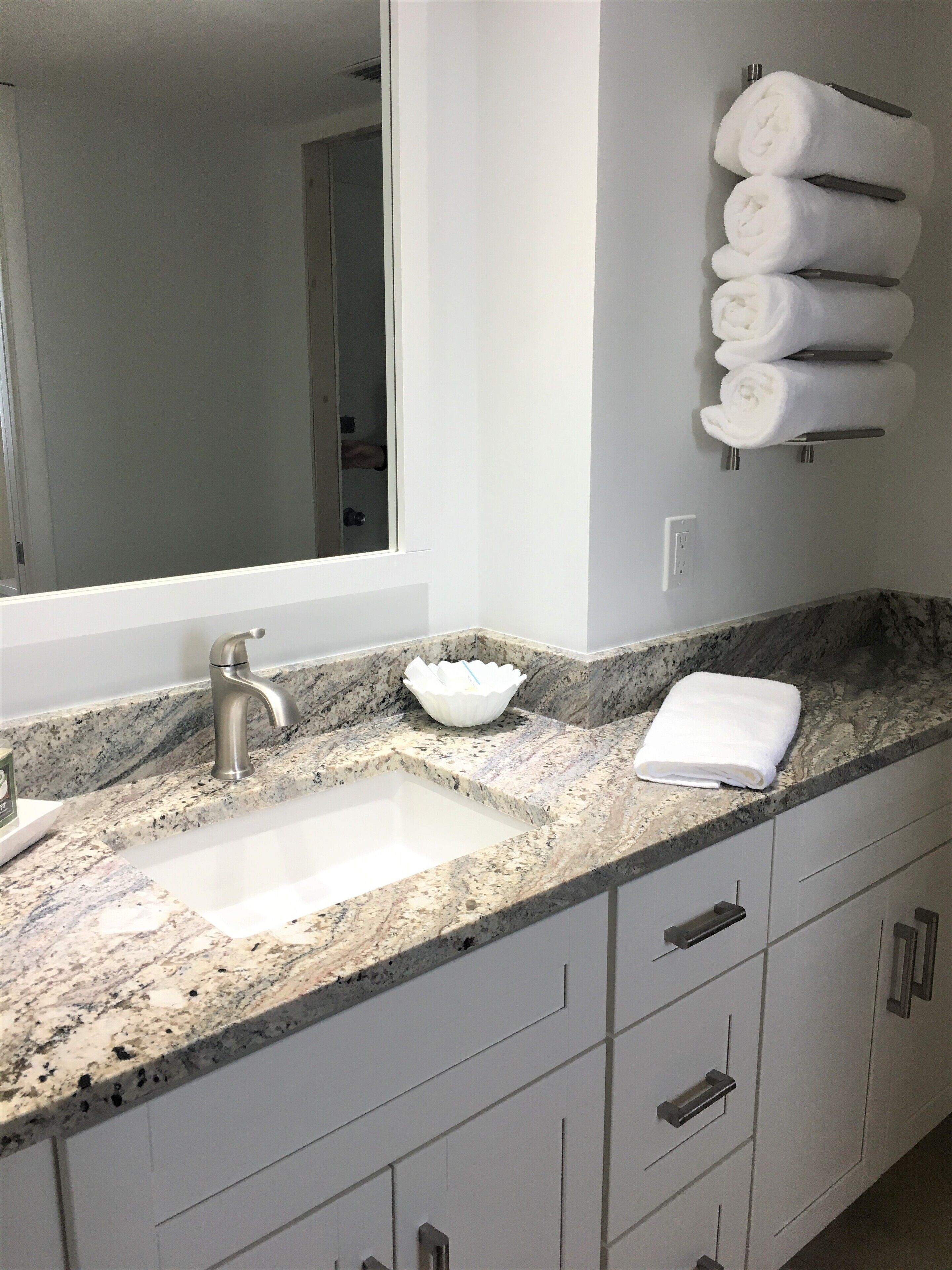 Une salle de bain dotée d’un comptoir en granit, d’un lavabo blanc, d’un miroir et d’un porte-serviettes avec des serviettes pliées.