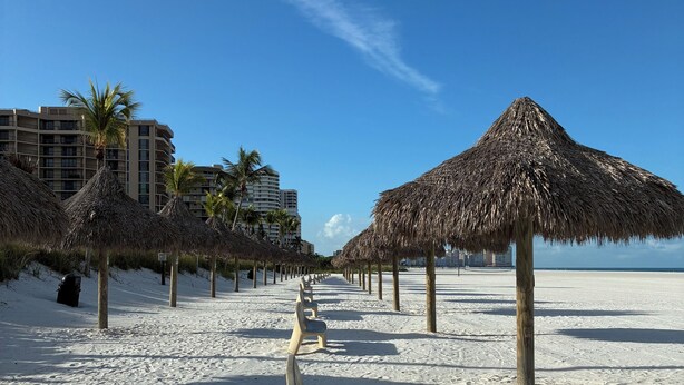 A beach with thatched umbrellas, a row of benches, and a hotel in the background. - Marco Island Riverside Condo - Comfort Meets Coastal Charm (Marco Island)