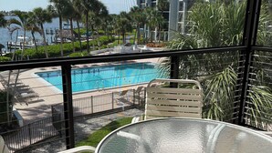 A balcony with a glass table and chairs overlooking a swimming pool, palm trees, and a building in the background. - Marco Island Riverside Condo - Comfort Meets Coastal Charm (Marco Island)