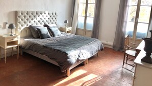 Ein Schlafzimmer mit einem gepolsterten Kopfteil, einem Bett mit grauer Bettdecke, zwei Nachttischen mit Lampen, einem Fenster mit Vorhängen und einer Pendelleuchte.