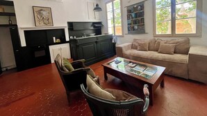 Ein Wohnzimmer mit Sofa, Couchtisch, Sessel, Fernseher, Bücherregal und einem Fenster mit Blick auf Bäume.