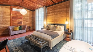A modern bedroom with a wooden ceiling, a large bed with multiple pillows, a bench, and a small table with a lamp. - Droombos (Windhoek)