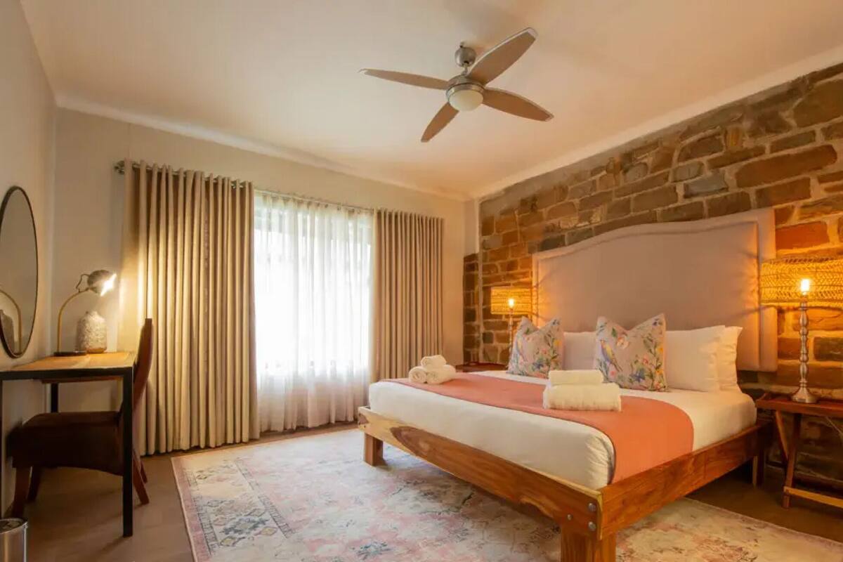 A bedroom with a stone wall, a bed with pillows, a ceiling fan, and a nightstand with a lamp.