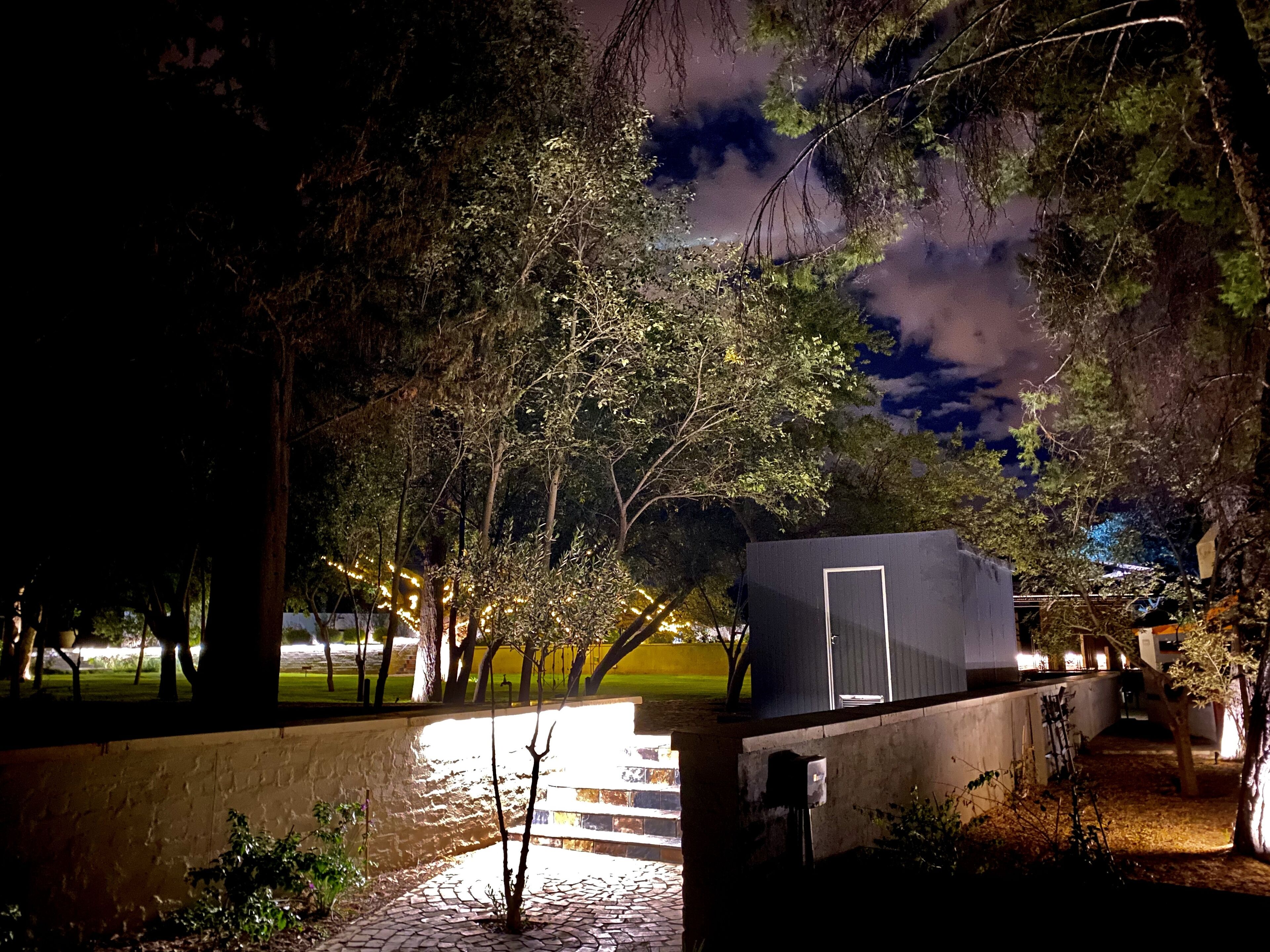 A small, dark-colored building with a door, surrounded by trees and a paved area.