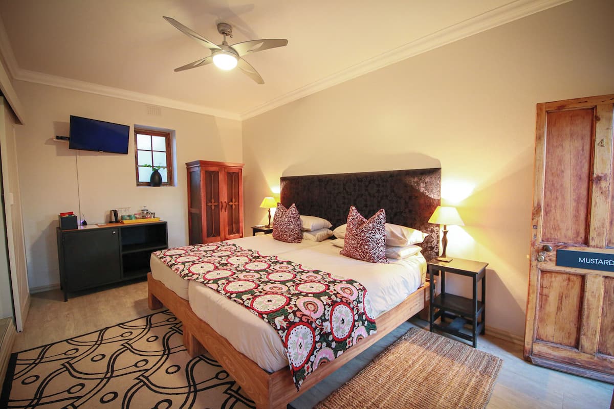 A bedroom with a bed, a ceiling fan, a TV, a window, a wooden wardrobe, and a door labeled "MUSTARD".