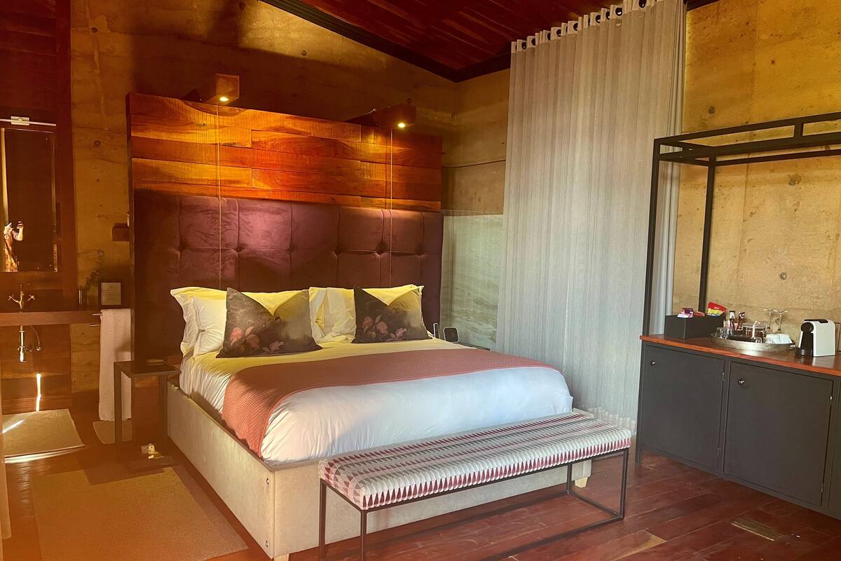A bedroom with a large bed, a bench, a wooden headboard, a mirror, and a small desk with a cup and a book.