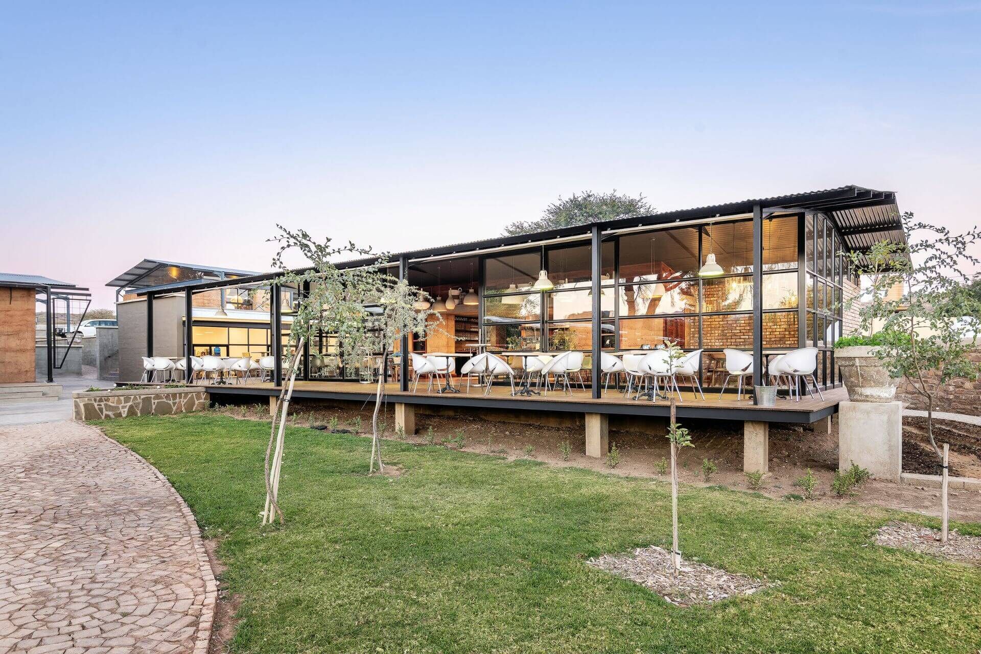 A modern outdoor dining area with glass walls, white chairs, and a brick building in the background.