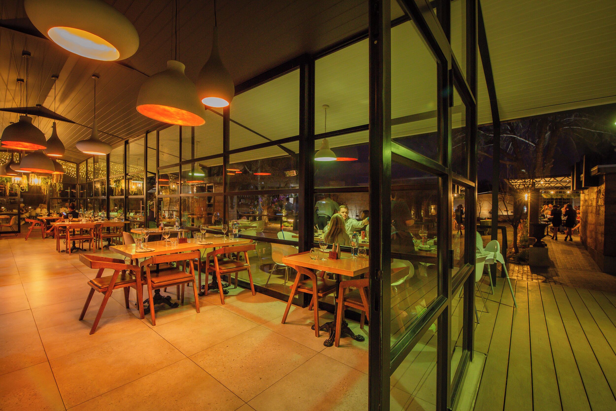 Een restaurant met houten tafels en stoelen, grote ramen en hangende lampen.